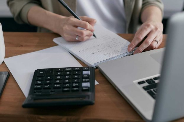 A woman writes financial calculations in a notebook, using a calculator and laptop at a wooden desk.